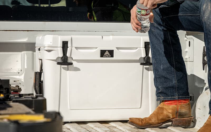 Person sitting in the back of a work truck drinking water from a white Apex A20 cooler