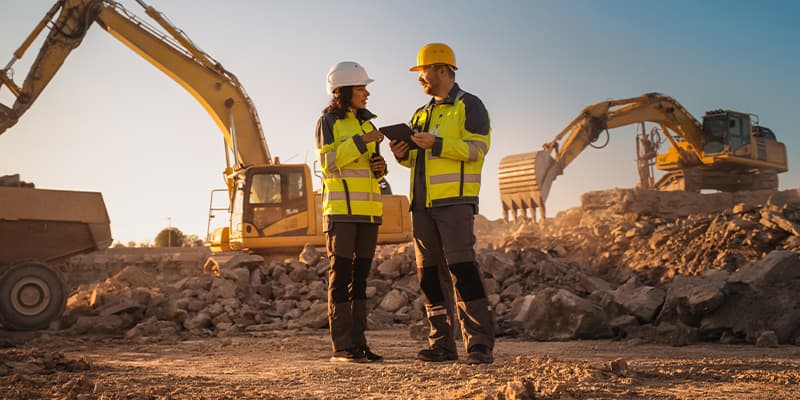 Two construction workers standing on a rocky construction site discussing plans with large excavators operating in the background.