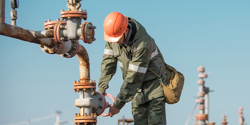 Worker in protective gear adjusting a valve on industrial piping at an outdoor facility